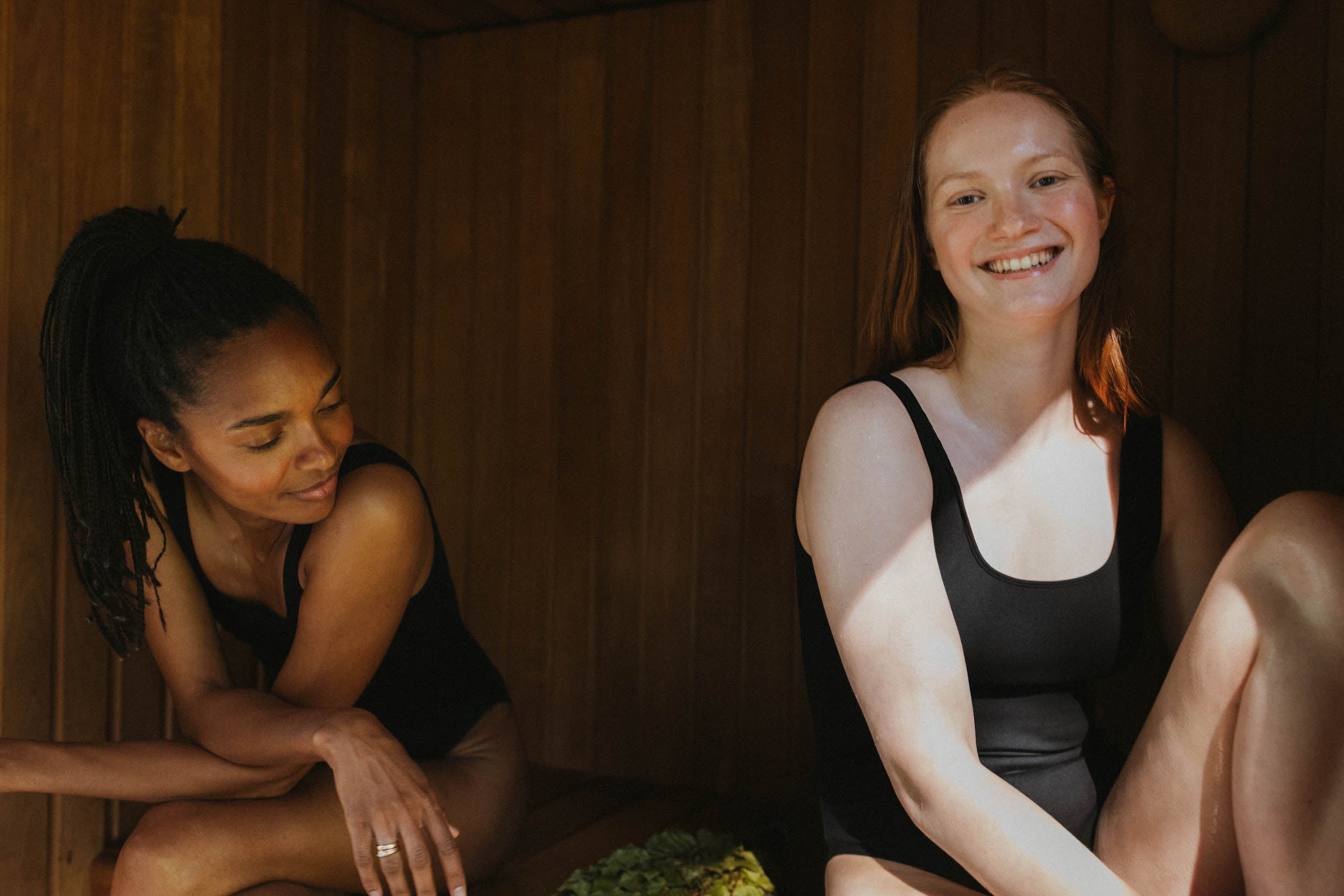 Two women sitting in a sauna, one smiling at the camera.